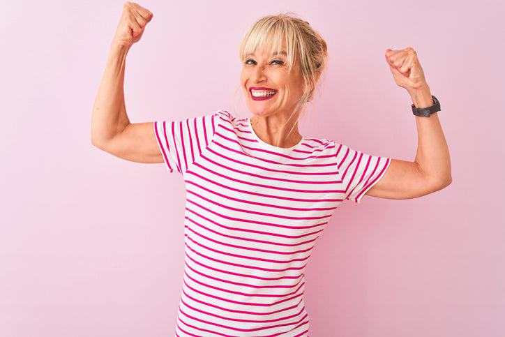 Glad moden kvinde i stribet T-shirt viser styrke med løftede arme og stort smil – symbol på energi, selvtillid og livsglæde.
