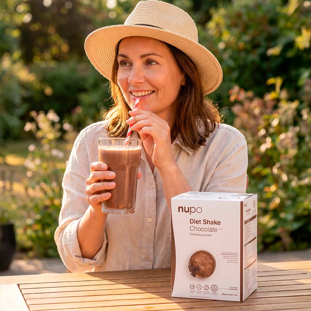 Woman enjoying a chocolate diet shake from Nupo on a sunny day, smiling with a straw in her glass.