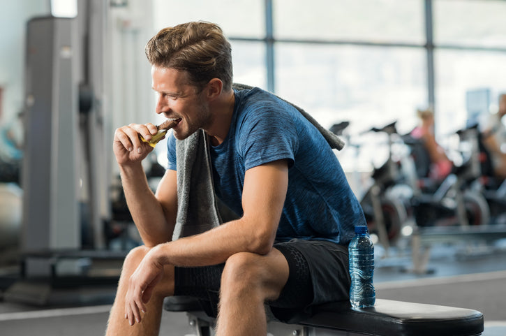 Ung mand i traeningstoej sidder paa baenk i fitnesscenter og spiser en proteinbar – afslappet pause efter workout med fokus paa restitution og sund livsstil.