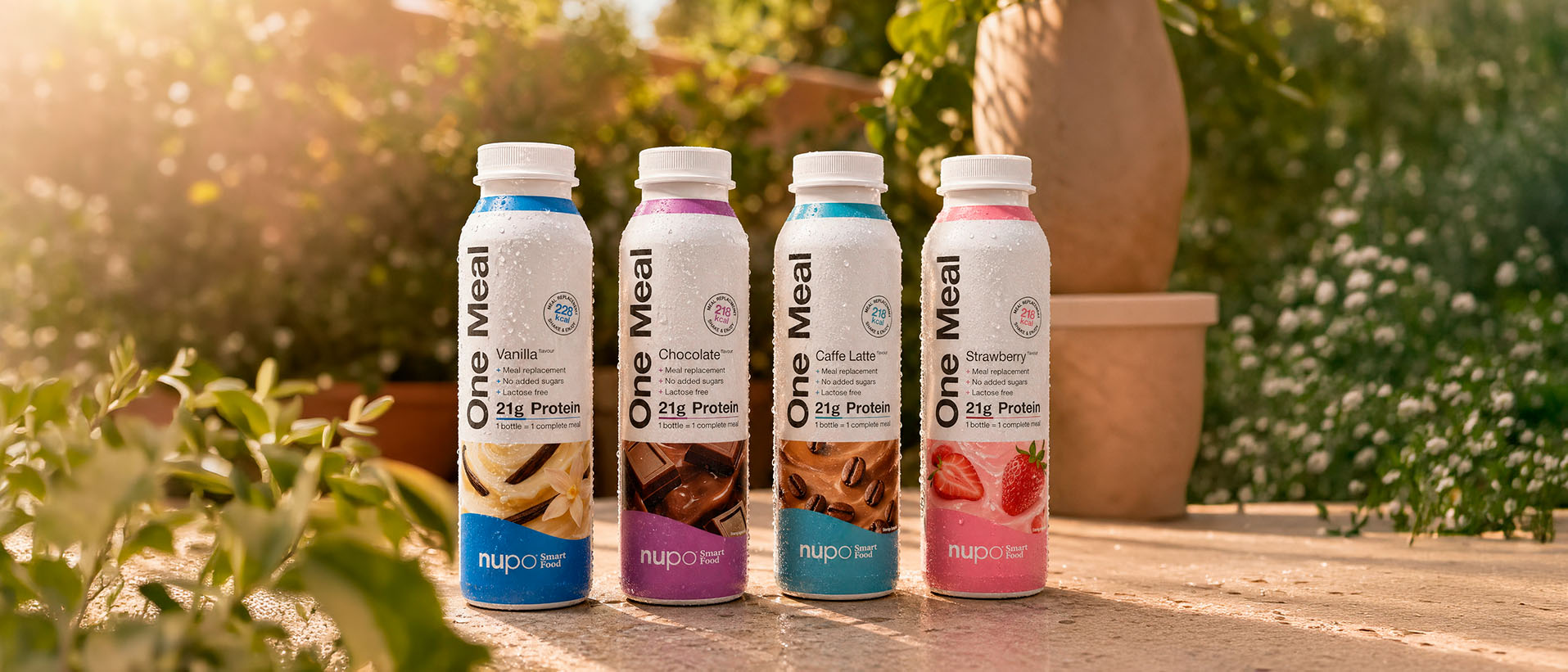 Four protein drink bottles in various flavors, placed outdoors with greenery and sunlight.