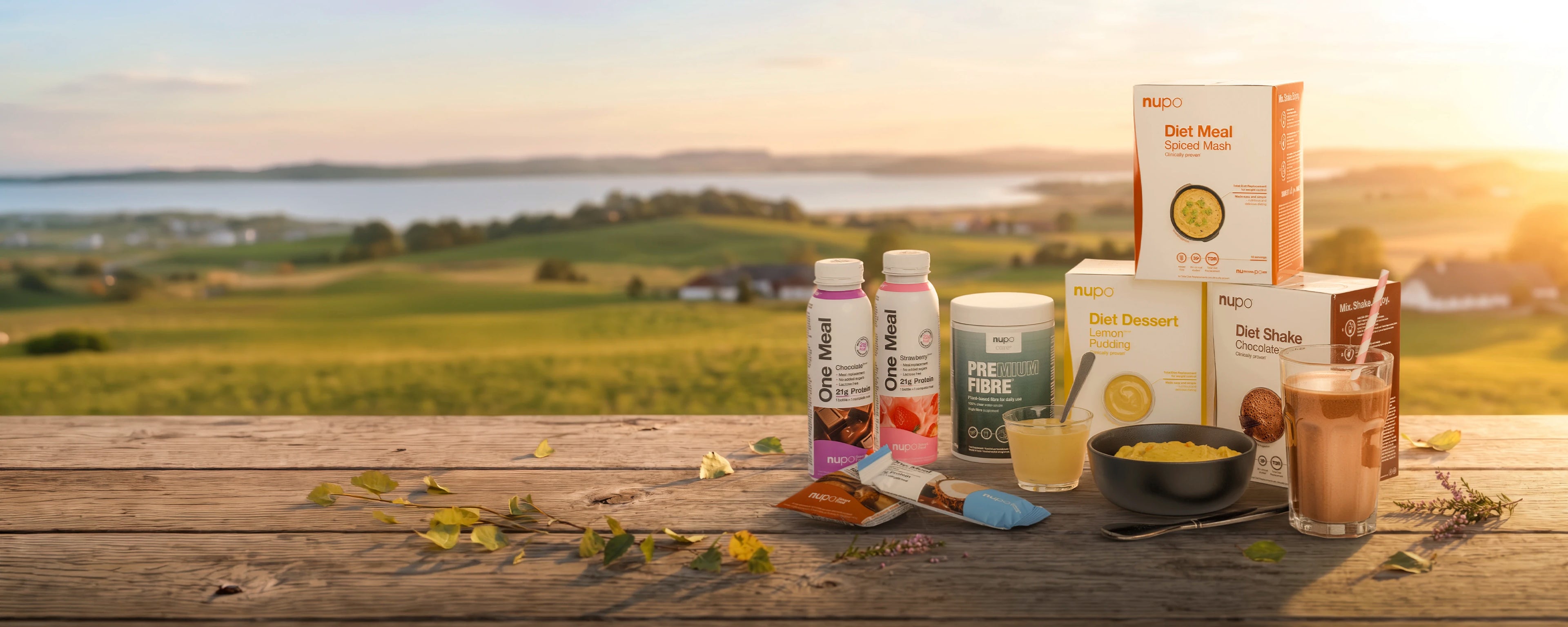 Assorted diet products displayed on a wooden table with a scenic countryside backdrop during sunset.